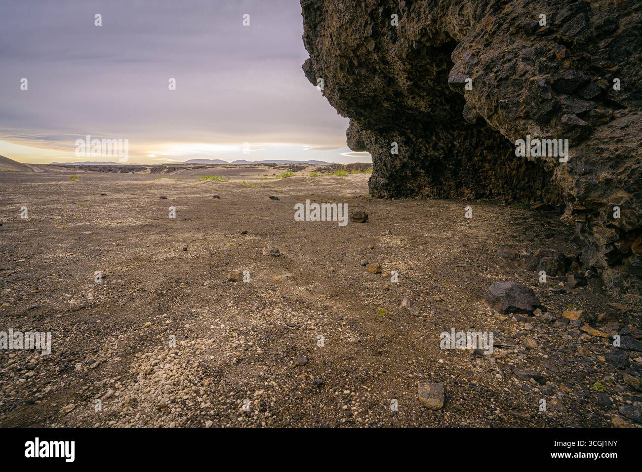 Dark volcanic cliffs rise over Iceland’s barren lava plains where scattered rocks and soft evening light create a stark cinematic landscape Stock Photo