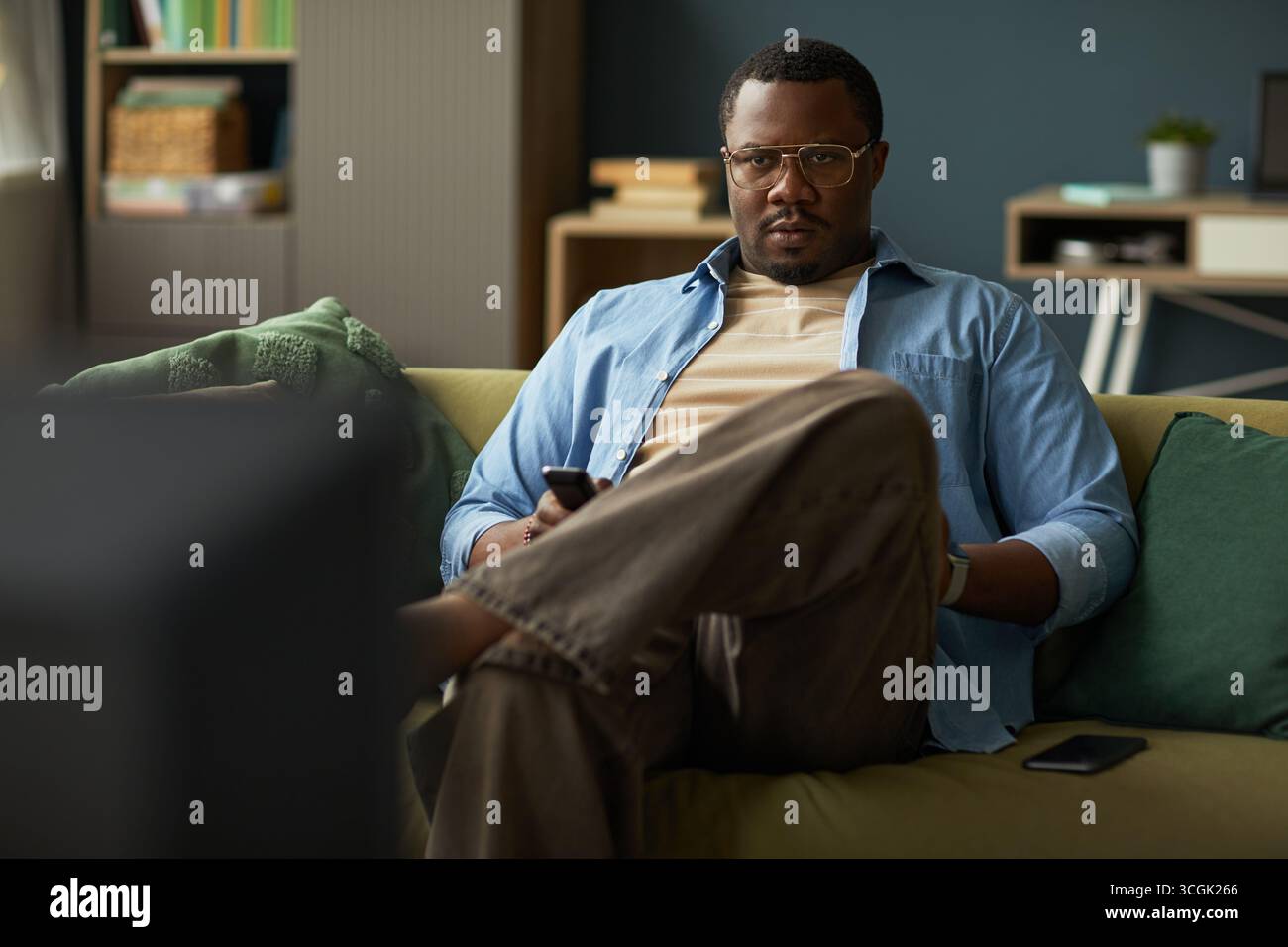 Portrait of Black man sitting on sofa holding remote control and watching television, wearing glasses and casual clothing, smartphone resting beside him on cushion Stock Photo