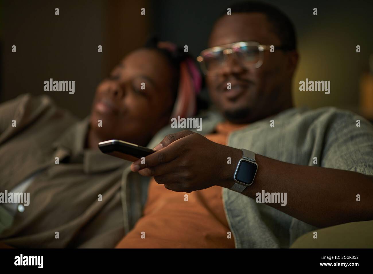 Black man holding remote control sitting next to Black woman relaxing on couch watching television together, both appearing comfortable and engaged in activity Stock Photo