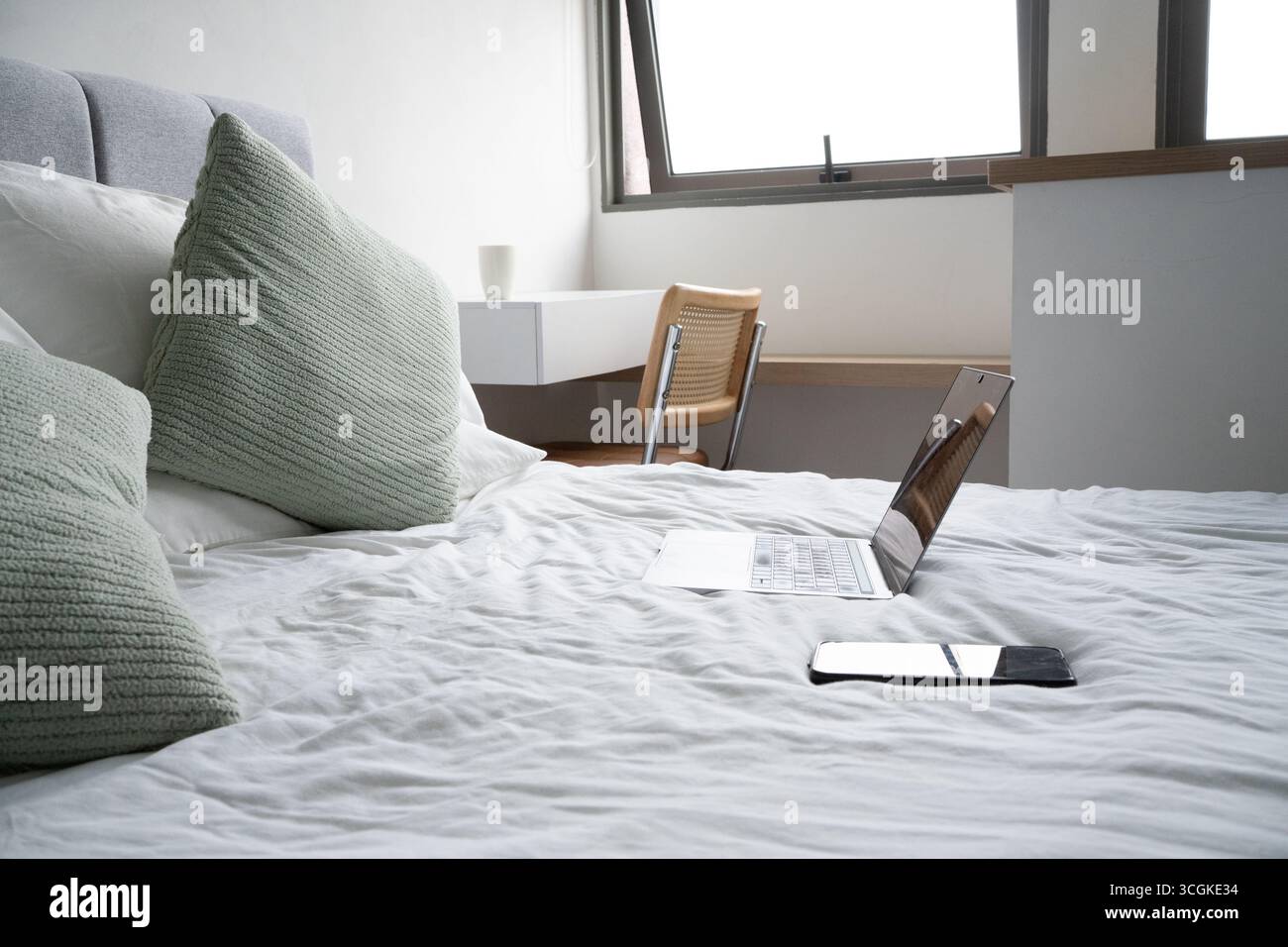Open laptop and smartphone lying on bed with crumpled sheets and green pillows, suggesting working from home or relaxing Stock Photo