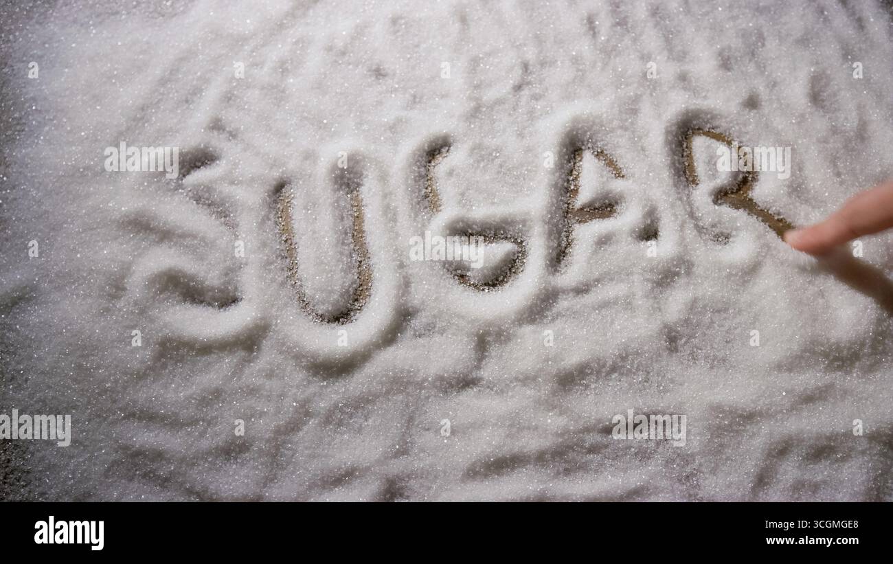 A hand writes the word sugar in a layer of white granulated sugar on a wooden table surface. Stock Photo
