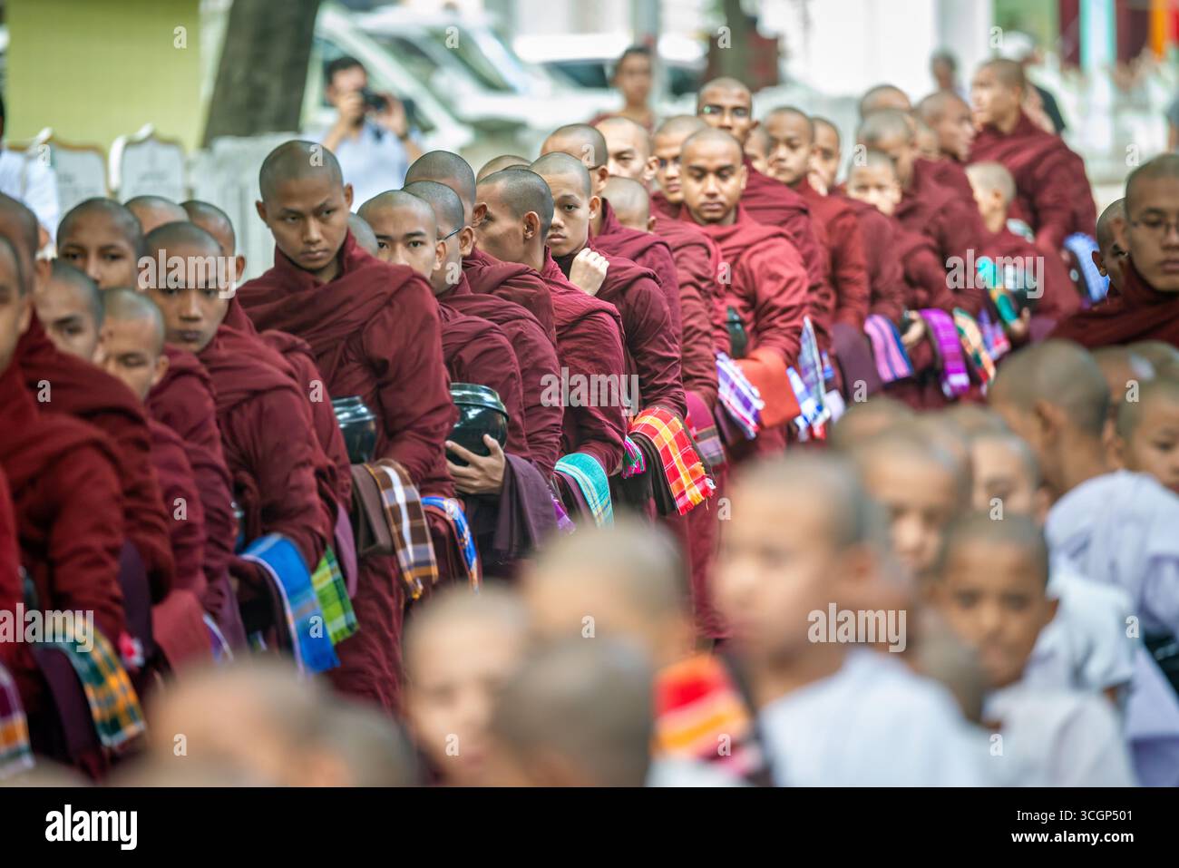 Mahagandayon Monastery Novices Lunchtime Procession Mandalay Myanmar // MANDALAY, MYANMAR — Novice Buddhist monks (samanera) line up with their alms bowls for the daily lunchtime meal procession at Mahagandayon Monastery. This daily ritual is a fundamental aspect of monastic life, where monks receive alms from lay devotees, symbolizing their reliance on the community. Mahagandayon Monastery (also known as Maha Gandayon Kyaung) is one of the largest and most prominent monastic colleges in Myanmar, home to thousands of monks and novices. It is a significant center for the study of Buddhist scrip Stock Photo