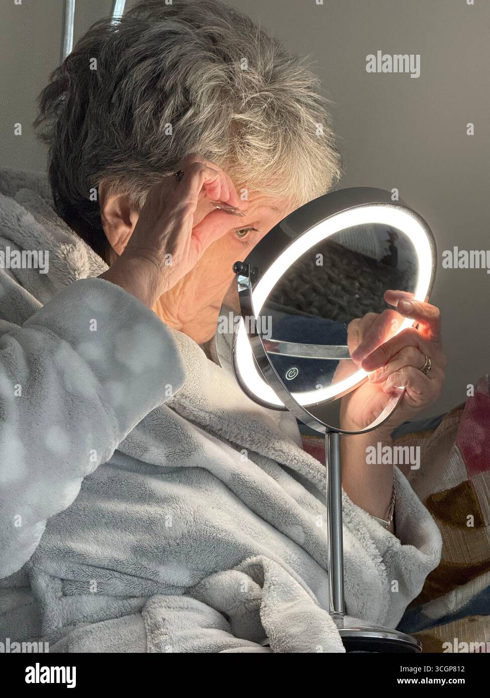 A mature lady in her bedroom and dressing gown plucking her eyebrows in a make up mirror - Smartphone Captured Stock Image