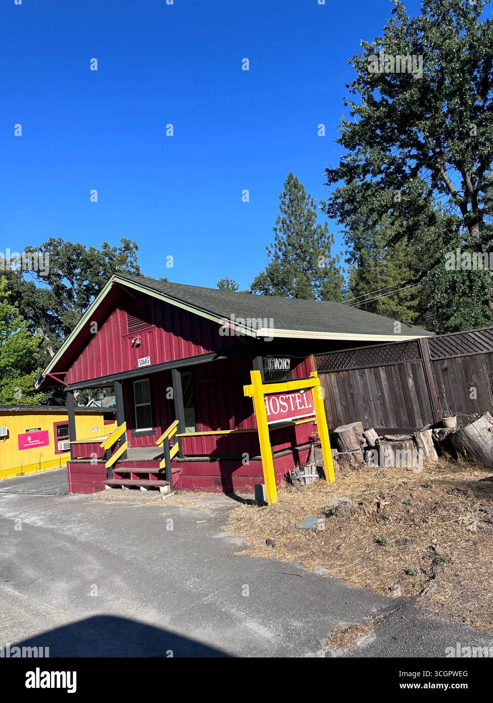 Red wooden hostel with yellow sign on a quiet street, surrounded by trees under a clear blue sky. - Smartphone Captured Stock Image