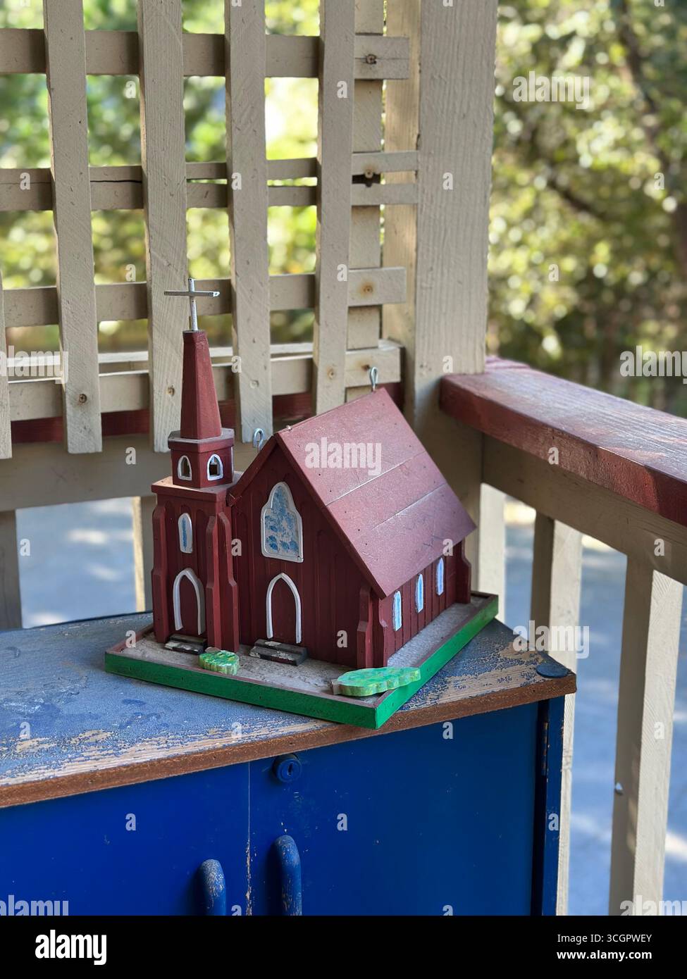 A handcrafted wooden model of a small red church with a steeple and painted details, placed on a shelf outdoors in natural daylight. - Smartphone Captured Stock Image
