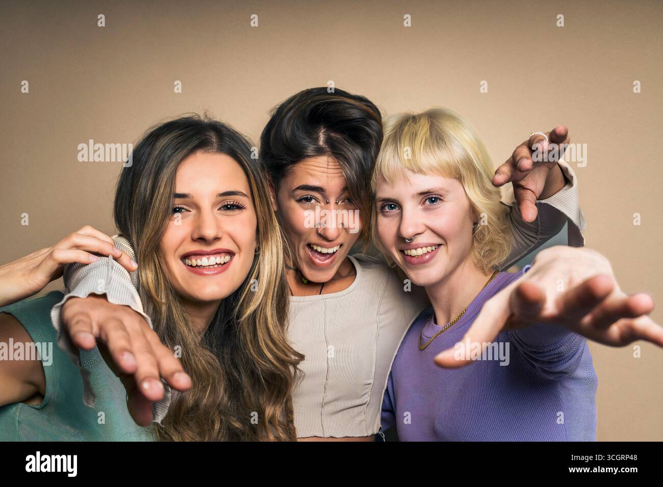 Playful studio portrait of three diverse and happy young women reaching their hands out to the camera in a fun and inviting gesture, as if to say 'wel Stock Photo