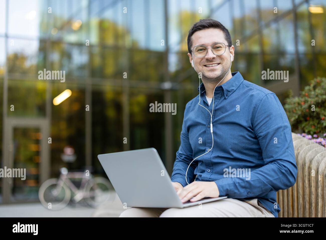 A cheerful individual in a blue shirt using a laptop outside a contemporary building. The setting emphasizes productivity, modern work environments, and a blend of technology with outdoor relaxation. Stock Photo