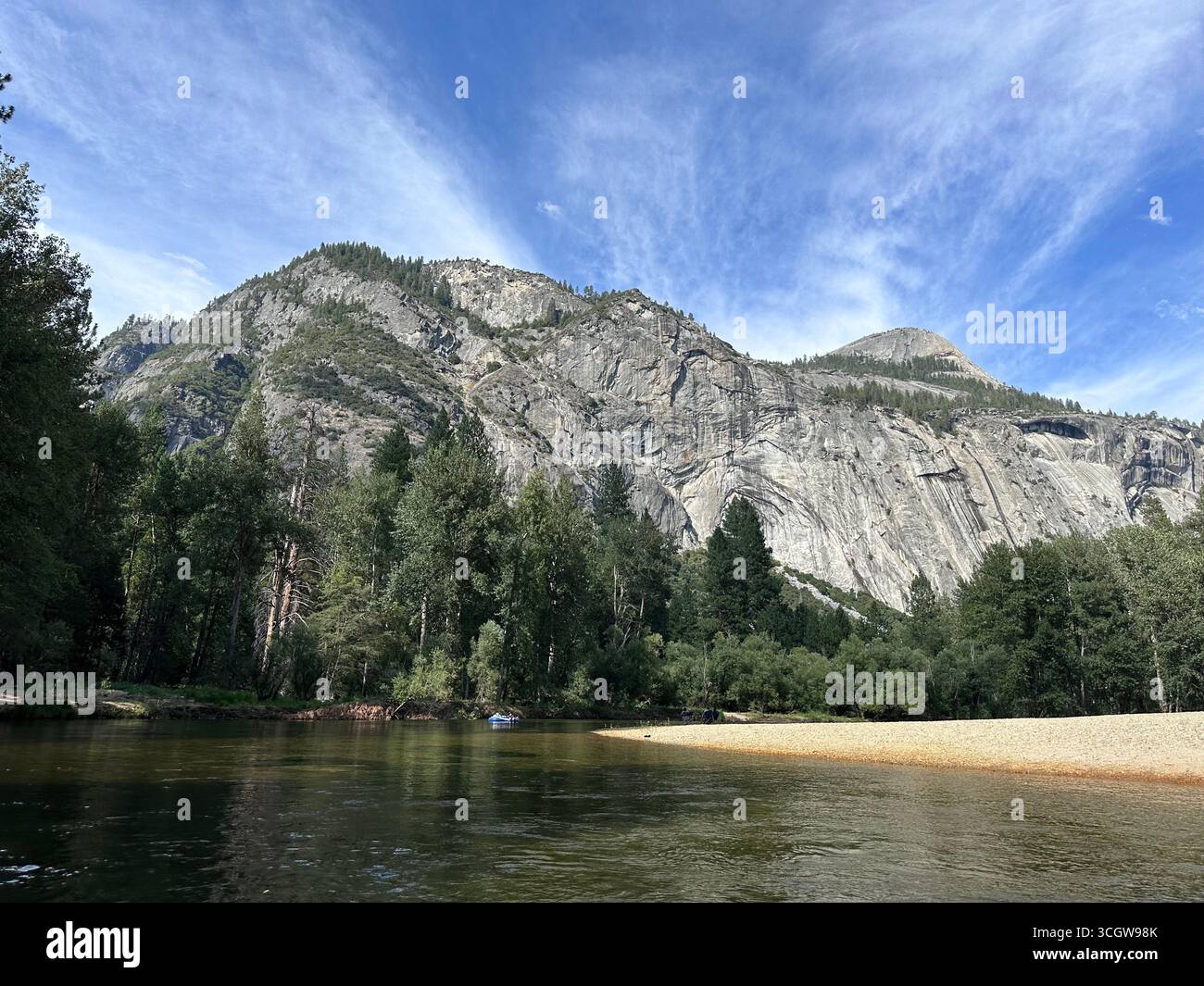 Scenic view of a rocky mountain with dense green forest and river foreground under a bright blue sky with wispy clouds. - Smartphone Captured Stock Image