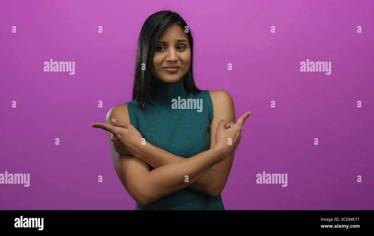 Woman expressing uncertainty against a vivid pink background, pointing with both hands, dressed in teal, displaying mixed emotions, isolated for conce Stock Photo