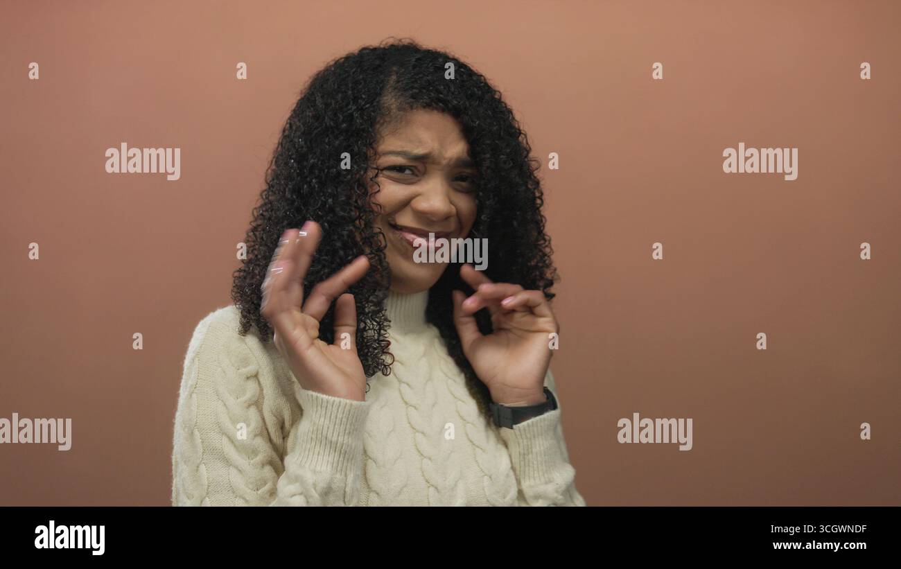 Recloiling Woman with curly hair raising hands in brown studio while recoiling ...