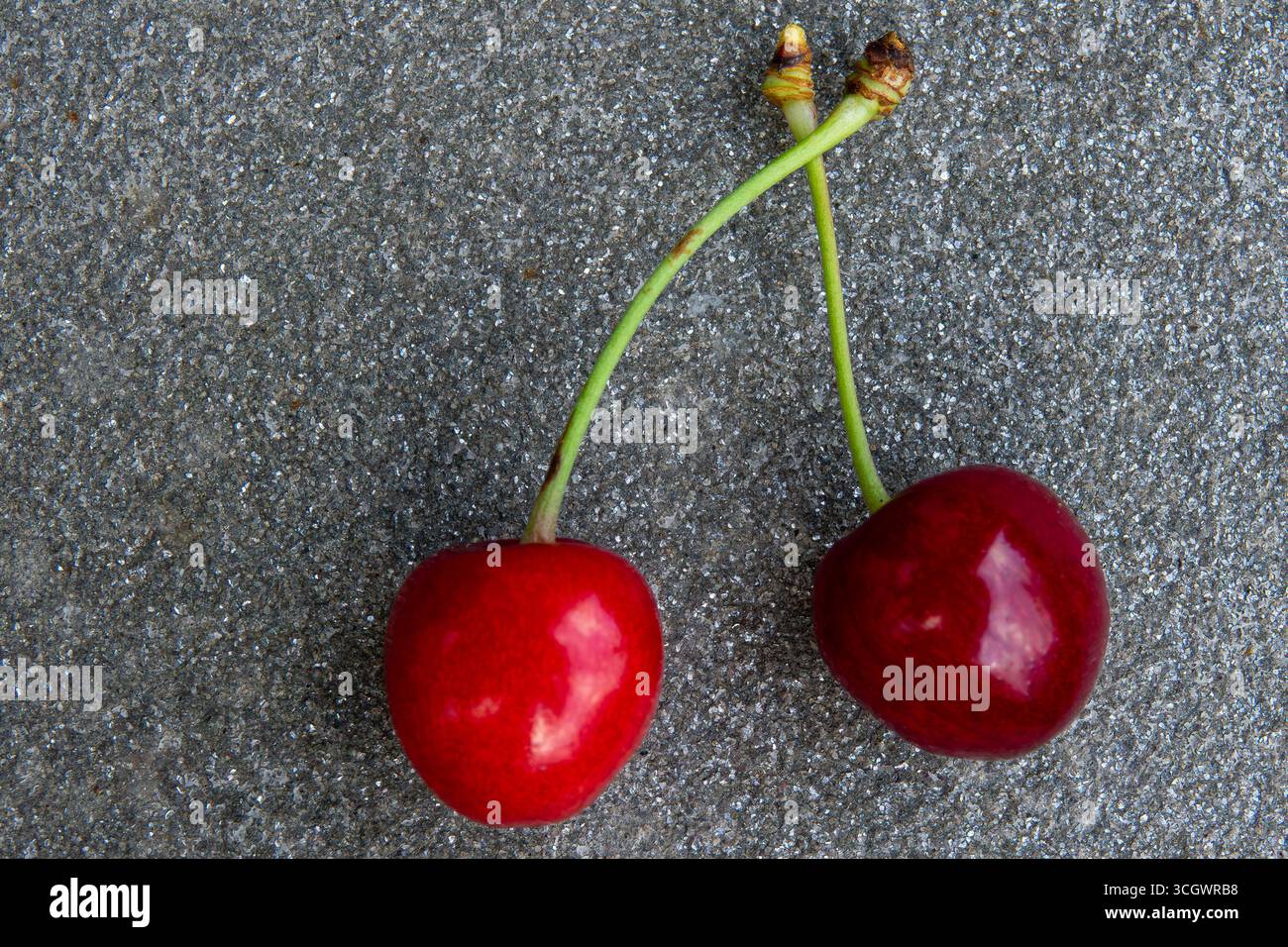 Two vibrant red cherries with stems positioned closely together on a textured gray stone surface, illuminated by soft lighting. Stock Photo