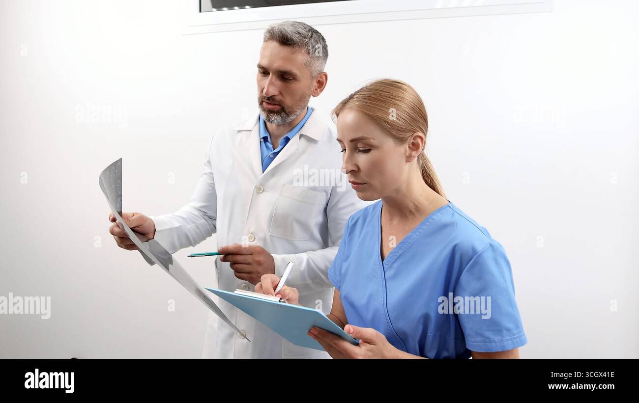 Doctor with MRI of patient in hands is standing in hospital with radiologic technologist and at looking in results of check-up Stock Photo