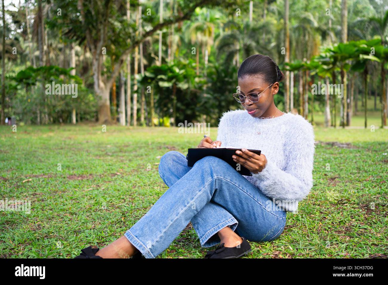Focused female student is taking notes on her notebook while sitting on the grass in a park, enjoying the fresh air and peaceful environment Stock Photo