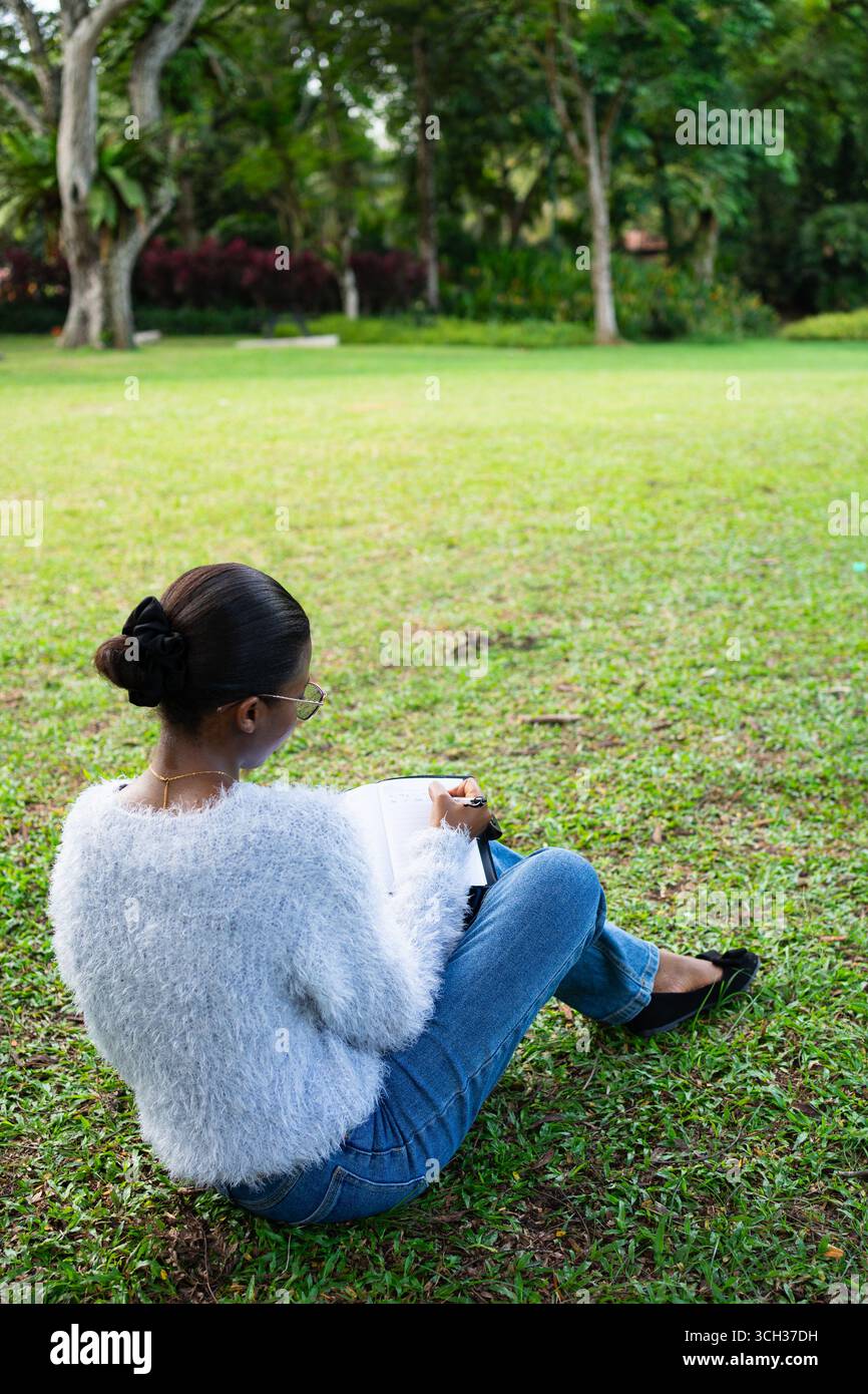 Student is taking notes sitting on grass in a park, enjoying her study time outdoors Stock Photo