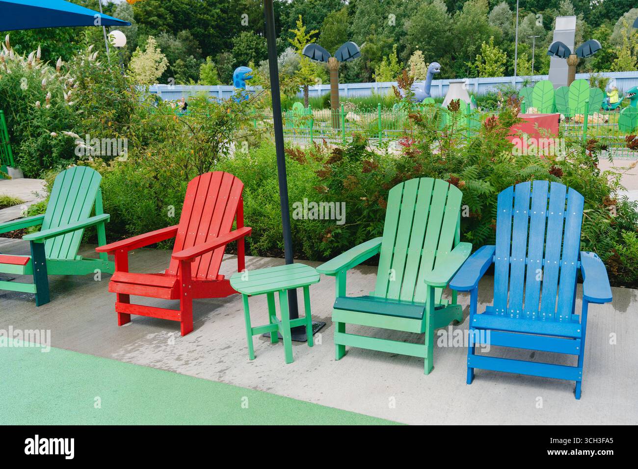 Vibrant red, green, and blue Adirondack chairs arranged in a park with lush greenery and a playground backdrop, creating a serene outdoor seating area Stock Photo