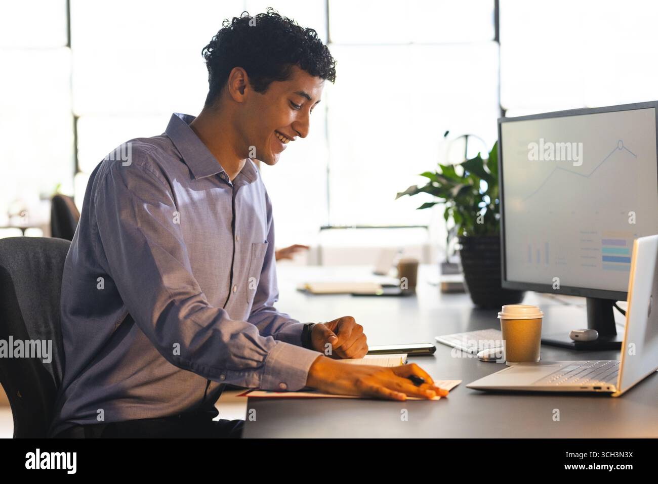 Smiling man at office desk analyzing data on computer, holding pen Stock Photo