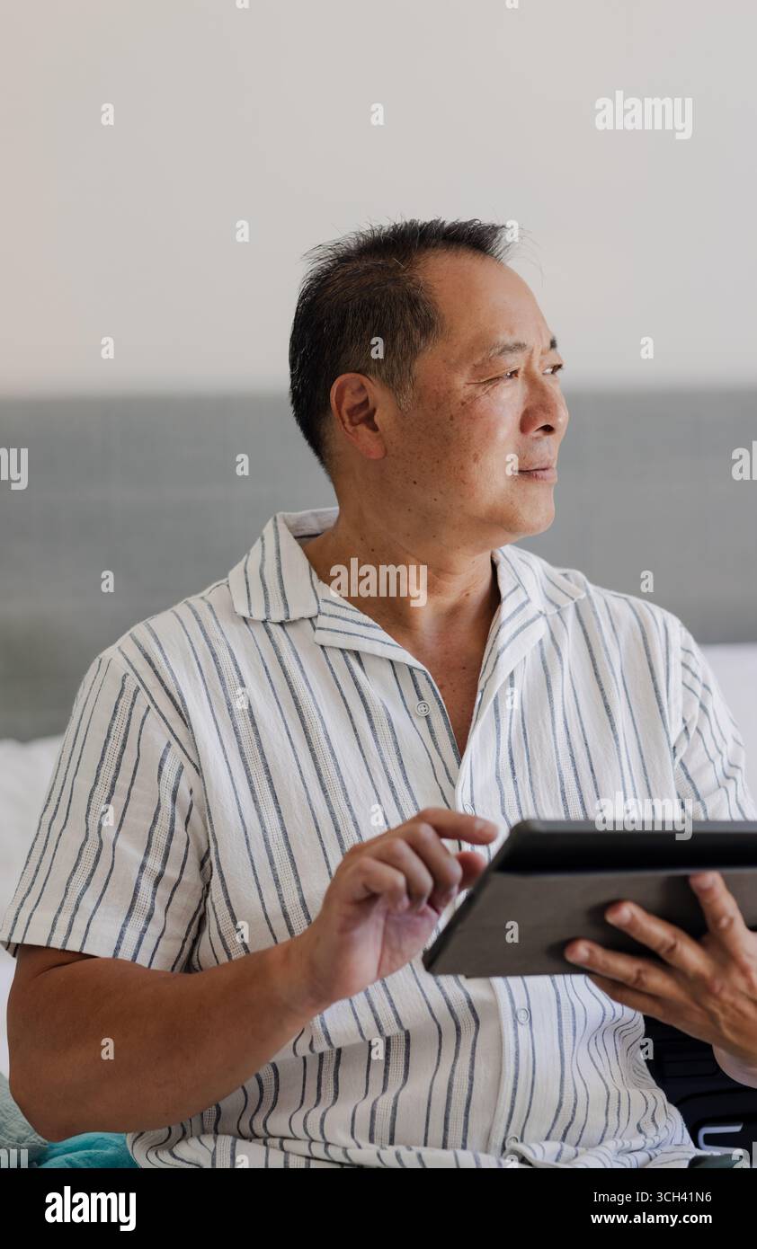 Senior man using tablet at home, looking thoughtfully out window Stock Photo