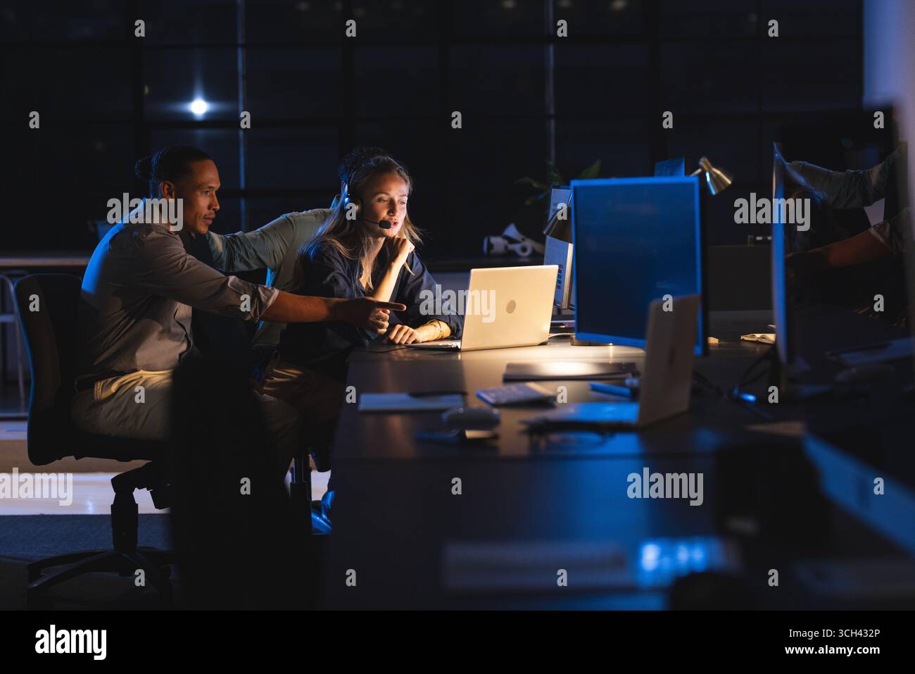 Colleagues collaborating on laptop in modern office during late-night work session Stock Photo