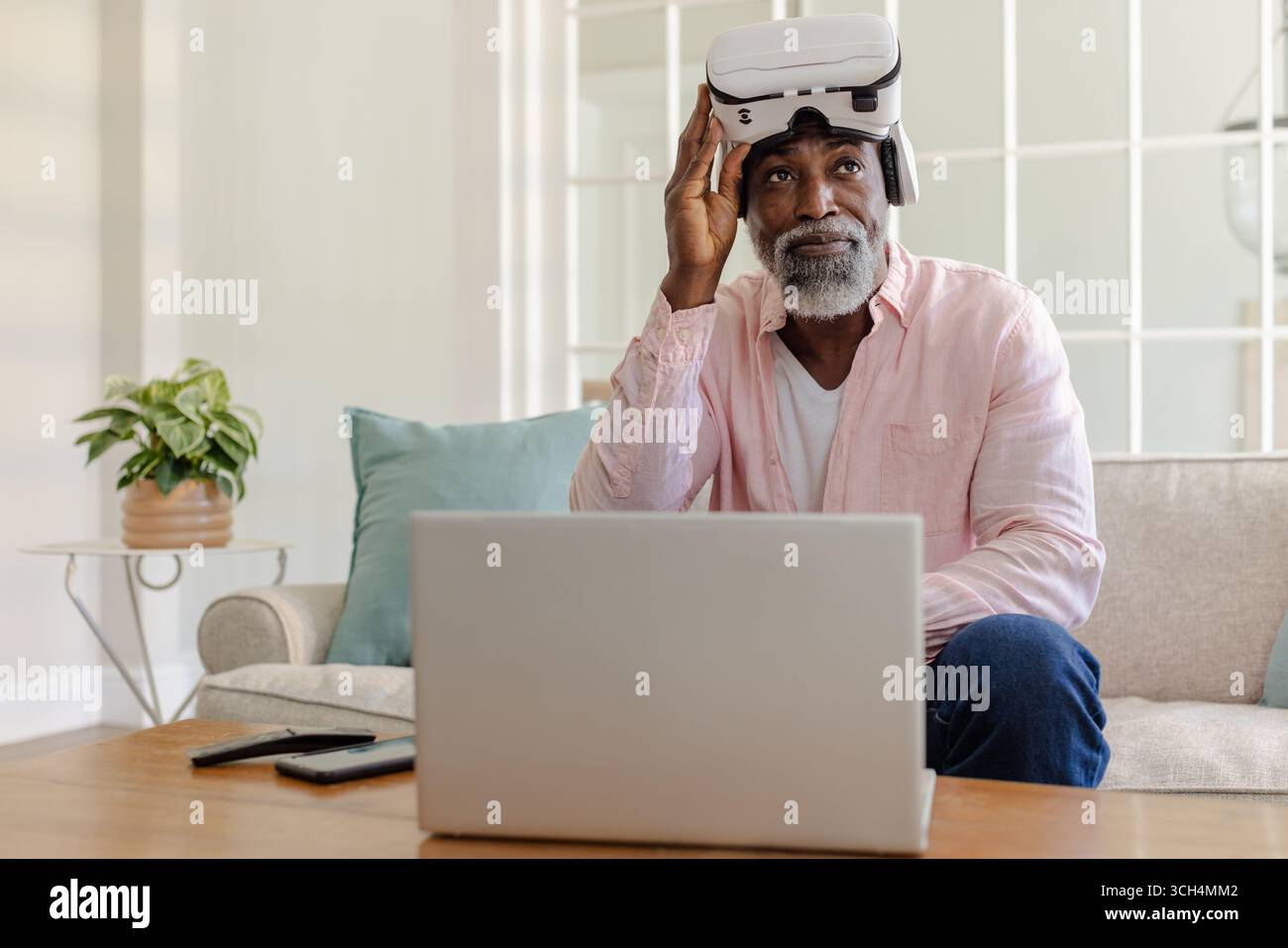 Senior man using VR headset and laptop at home, exploring virtual reality Stock Photo