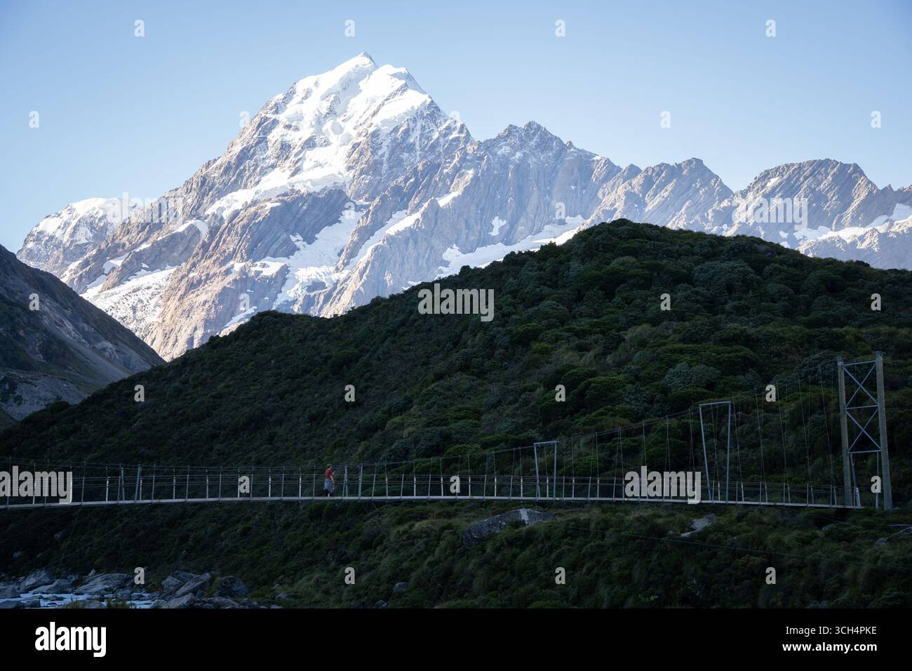 Tourist walking a swing bridge with huge snowy mountain in backdrop, Mt Cook, New Zealand Stock Photo