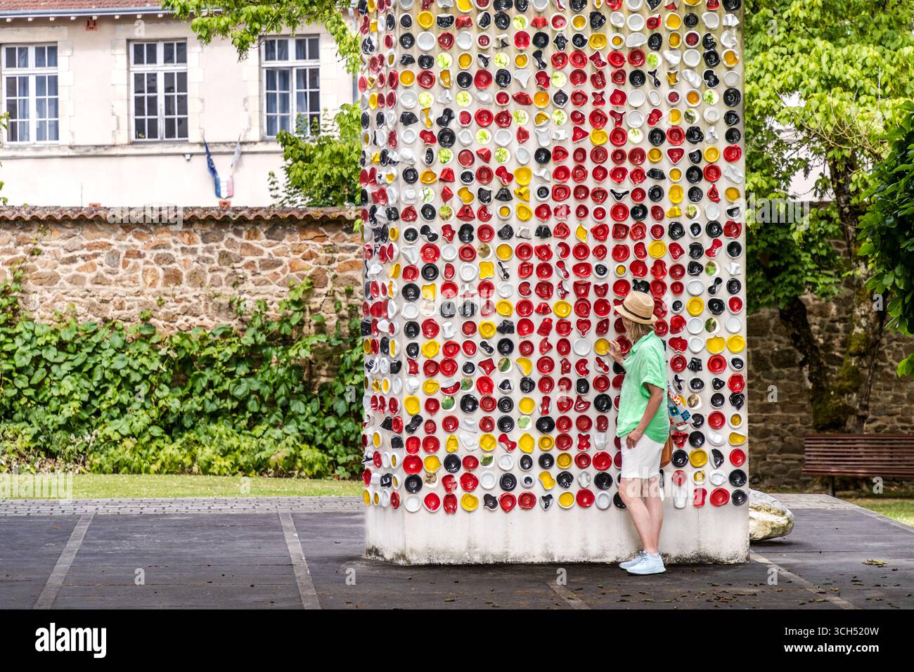 The art installation "Une Suite" by the artists Haguiko and Jean-Pierre Viot stands in front of the Musée National Adrien Dubouché on Place Winston Churchill in Limoges, Haute-Vienne, Nouvelle-Aquitaine, France. It consists of coloured, partly deformed porcelain bowls suspended from a tall, white concrete stele. In total, the work comprises around 600 porcelain bowls in red, yellow, black and white. Journalist Angela Berg, wearing a straw hat and a green blouse, stands in front of the stele and touches the bowls. The museum acquired the sculpture under the "1% artistique" public art scheme and Stock Photo