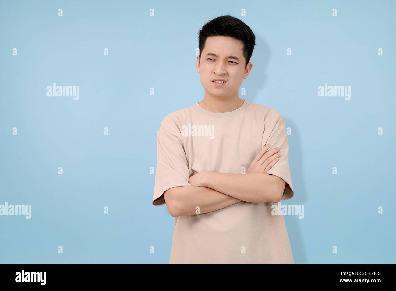 young man shows concerned expression with crossed arms against solid blue background Stock Photo