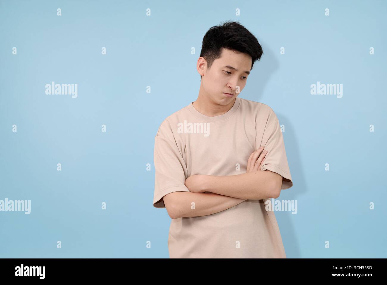 young man shows concerned expression with crossed arms against solid blue background Stock Photo