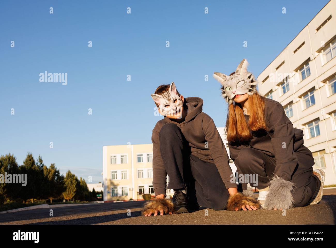 Two people wearing cat masks and furry gloves posing outdoors in costume under clear sky near school building Stock Photo
