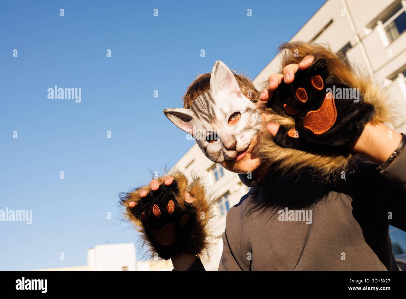 Creative street portrait of person wearing cat mask and furry gloves posing playfully in urban sunlight setting Stock Photo