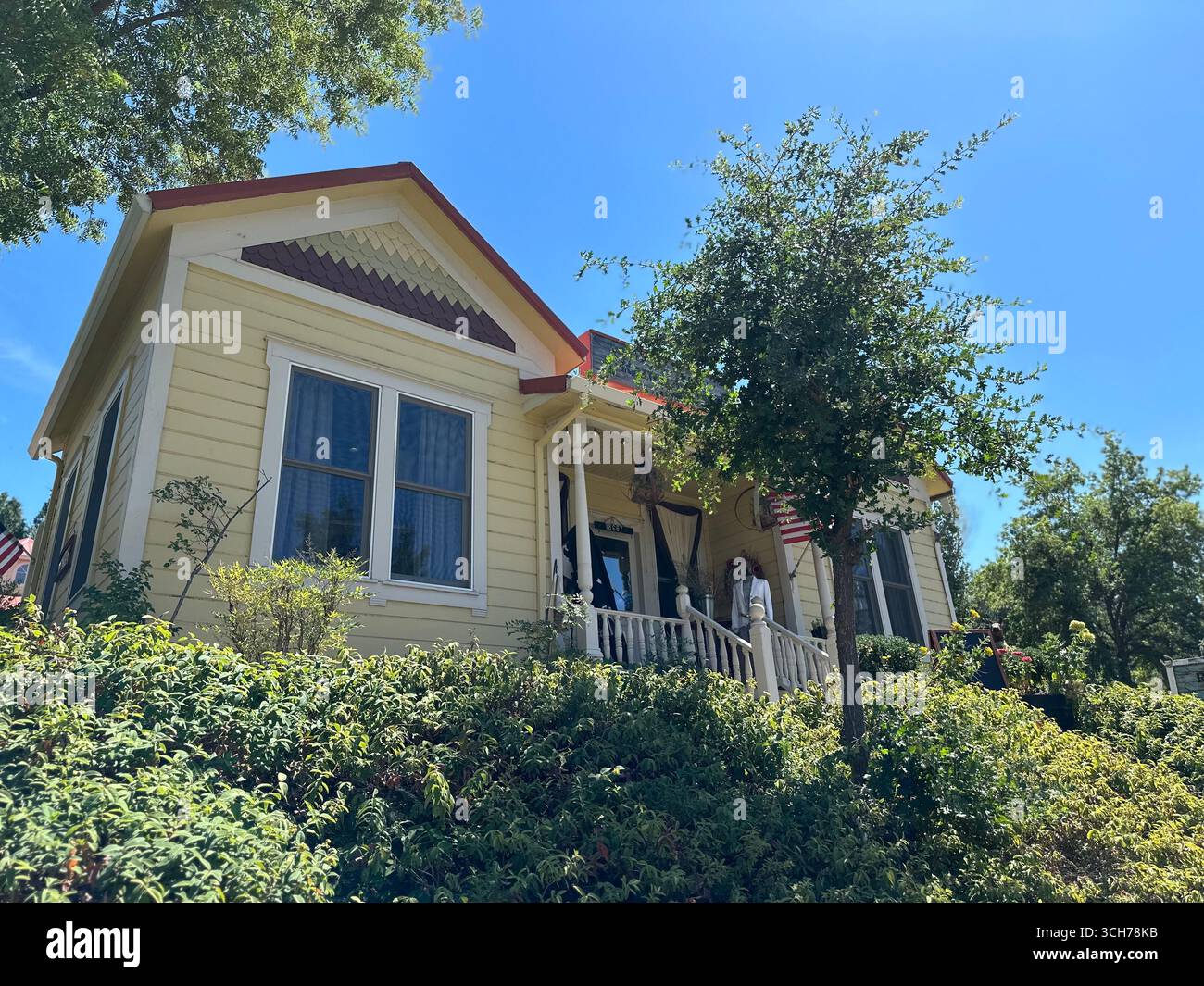 A yellow wooden house with front porch, American flag, and front garden plants, photographed from below under a clear blue sky. - Smartphone Captured Stock Image