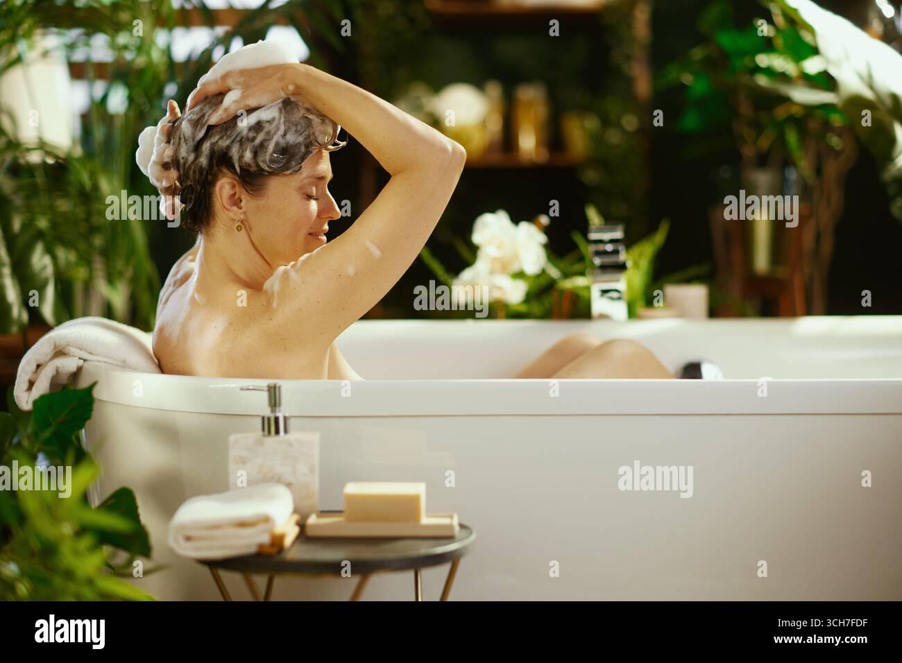 A mature woman with soapy hair relaxes in a bright white bathtub, eyes closed, enjoying a peaceful hair wash. Surrounded by lush green plants, this sc Stock Photo