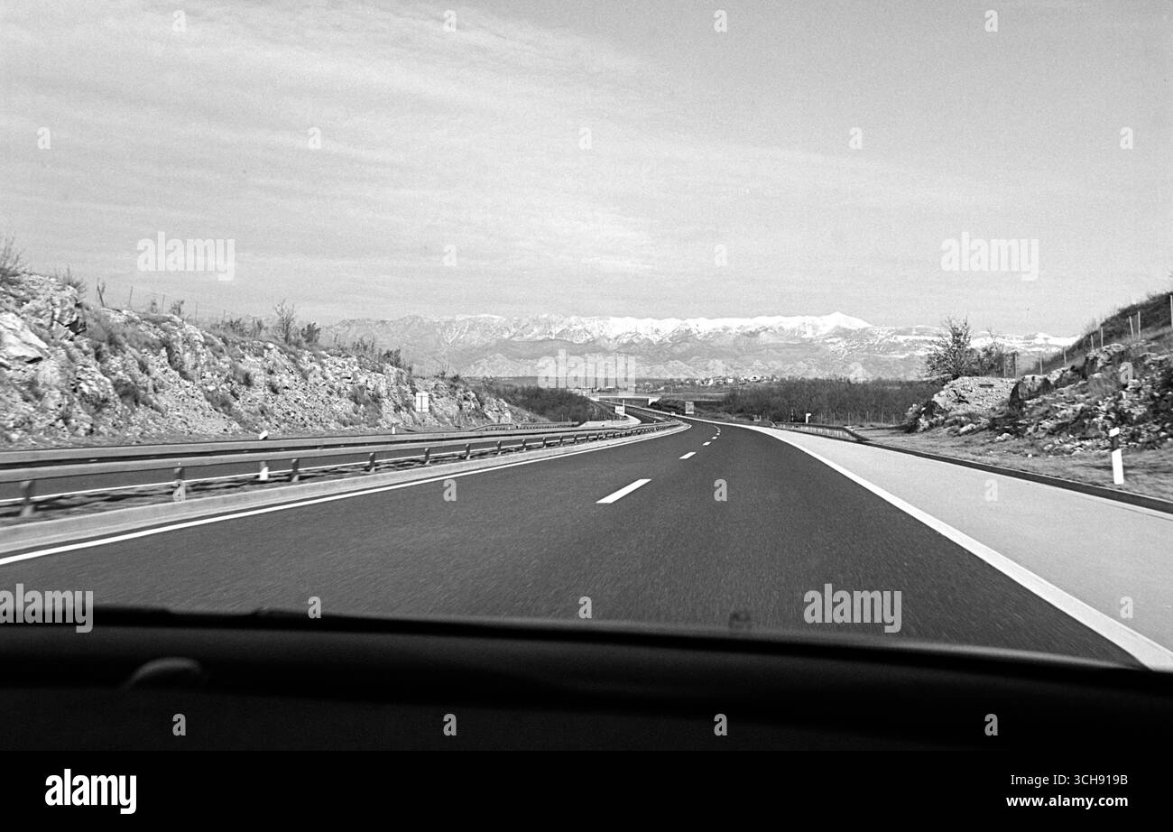 Black and white POV photo taken from the passenger seat of a moving car, showing an open highway stretching toward the distant Velebit mountain range. Stock Photo