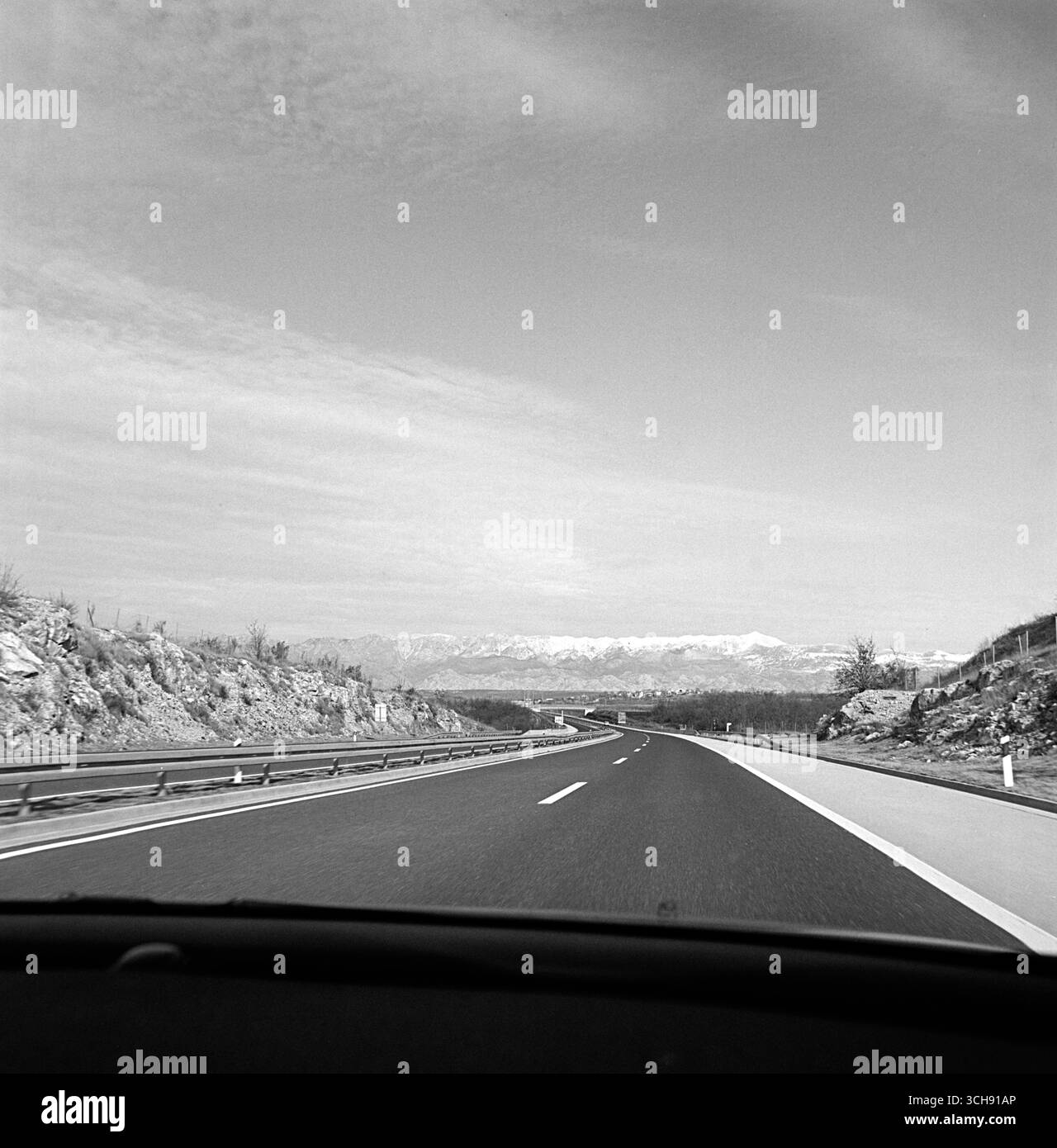 Black and white POV photo taken from the passenger seat of a moving car, showing an open highway stretching toward the distant Velebit mountain range. Stock Photo