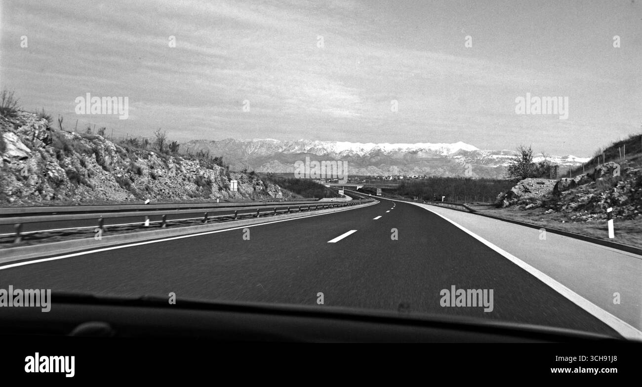 Black and white POV photo taken from the passenger seat of a moving car, showing an open highway stretching toward the distant Velebit mountain range. Stock Photo