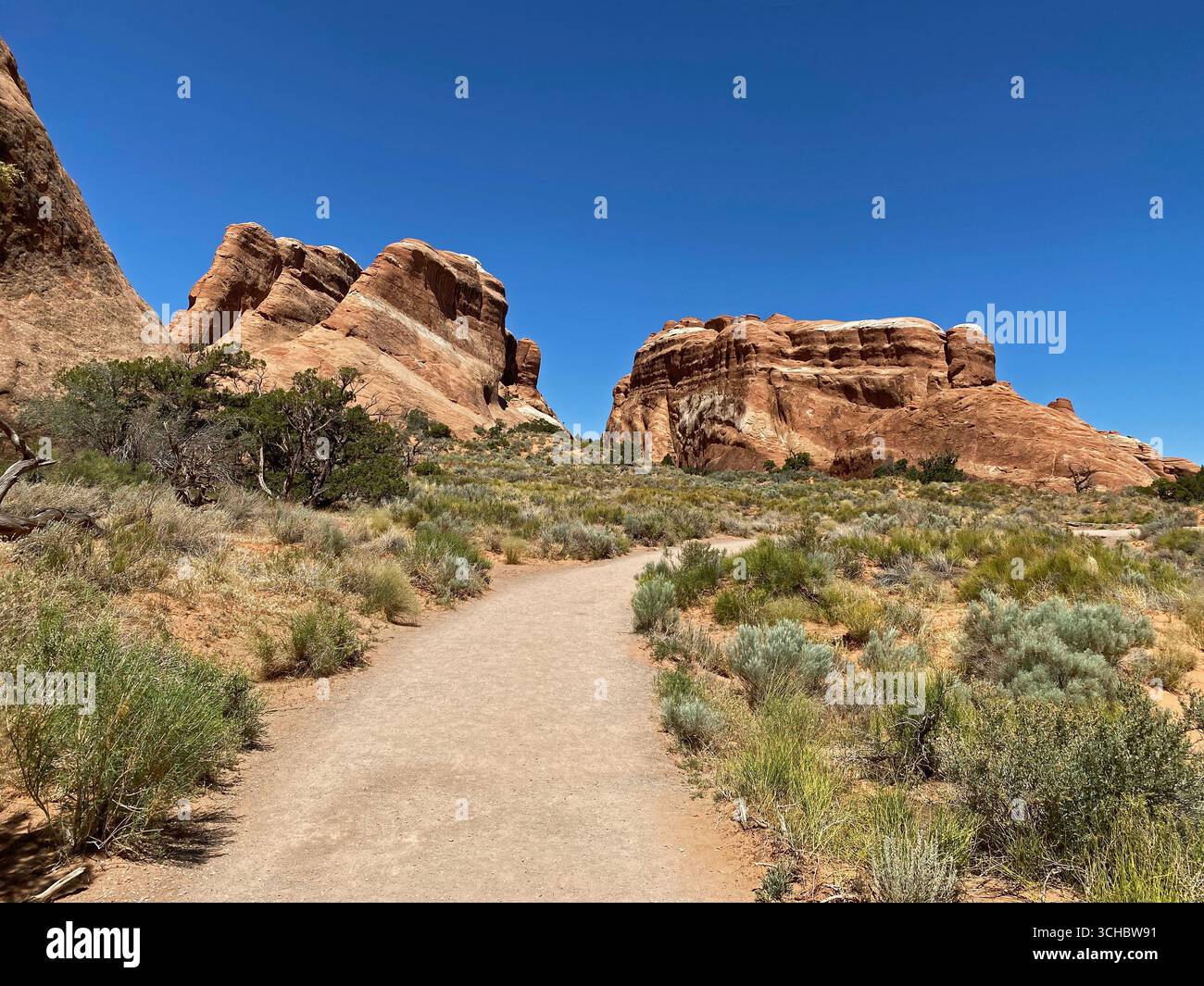 A trail leading through scrub brush and past large rock formations on the Landscape Arch Trail in Arches National Park, Moab, Utah, USA - Smartphone Captured Stock Image