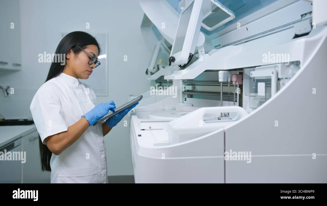 A laboratory technician in white attire uses a tablet to analyze data in a modern lab environment. Stock Photo