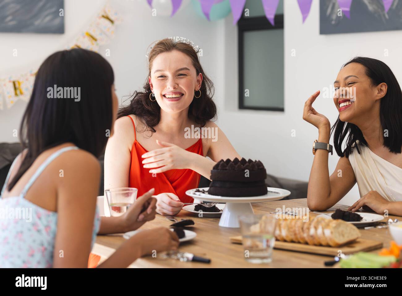 Diverse female friends celebrating at home, enjoying cake and laughter around festive table Stock Photo