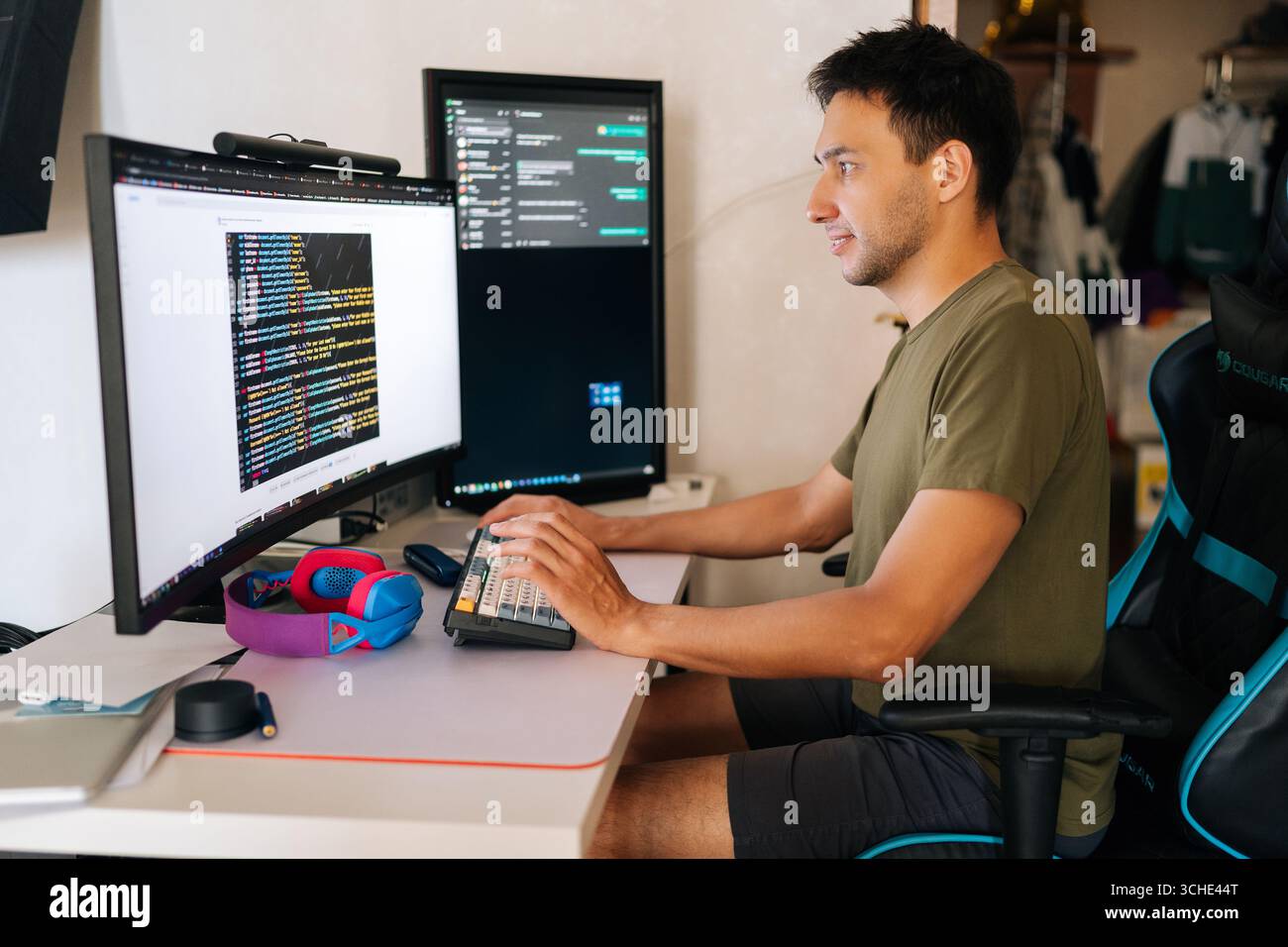 Focused software developer writing code at home office desk, surrounded by dual monitors displaying lines of code and communication platforms in Stock Photo