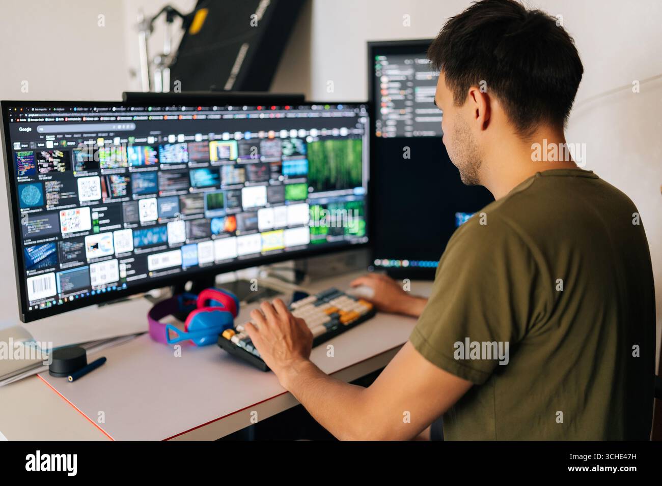 Back view of software developer male writing code at home office desk, surrounded by dual monitors displaying lines of code and communication Stock Photo