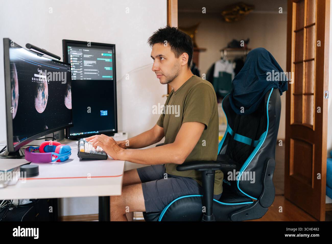 Successful programmer developer writing code at home office desk, surrounded by dual monitors displaying lines of code and communication platforms in Stock Photo