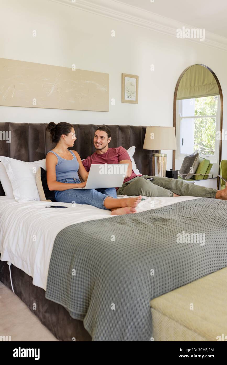 Lounging couple chatting over laptop on bed in modern bedroom, with decorative pillows Stock Photo