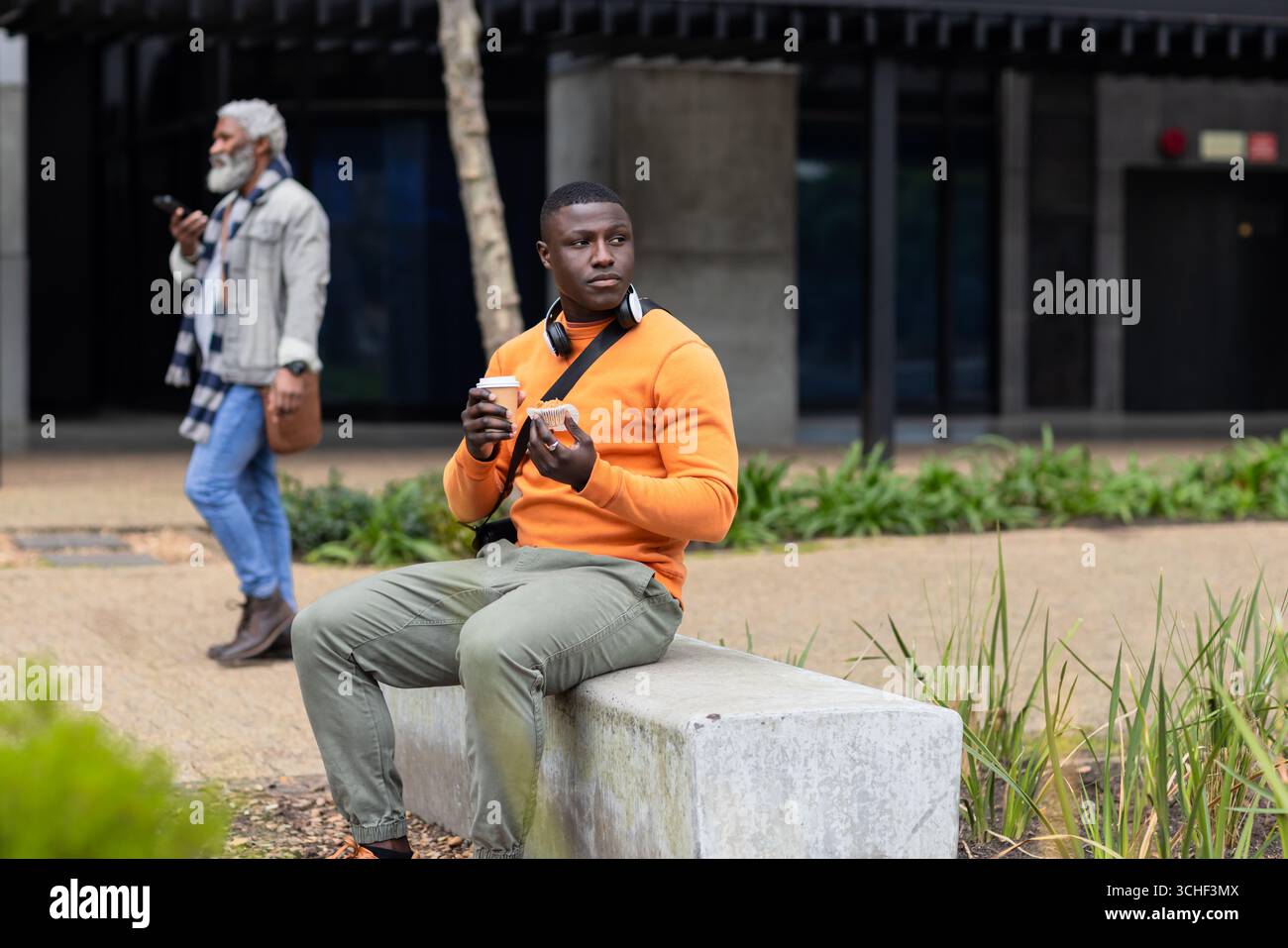 African american man wearing orange sweater sitting on bench in plaza holding coffee and pastry Stock Photo
