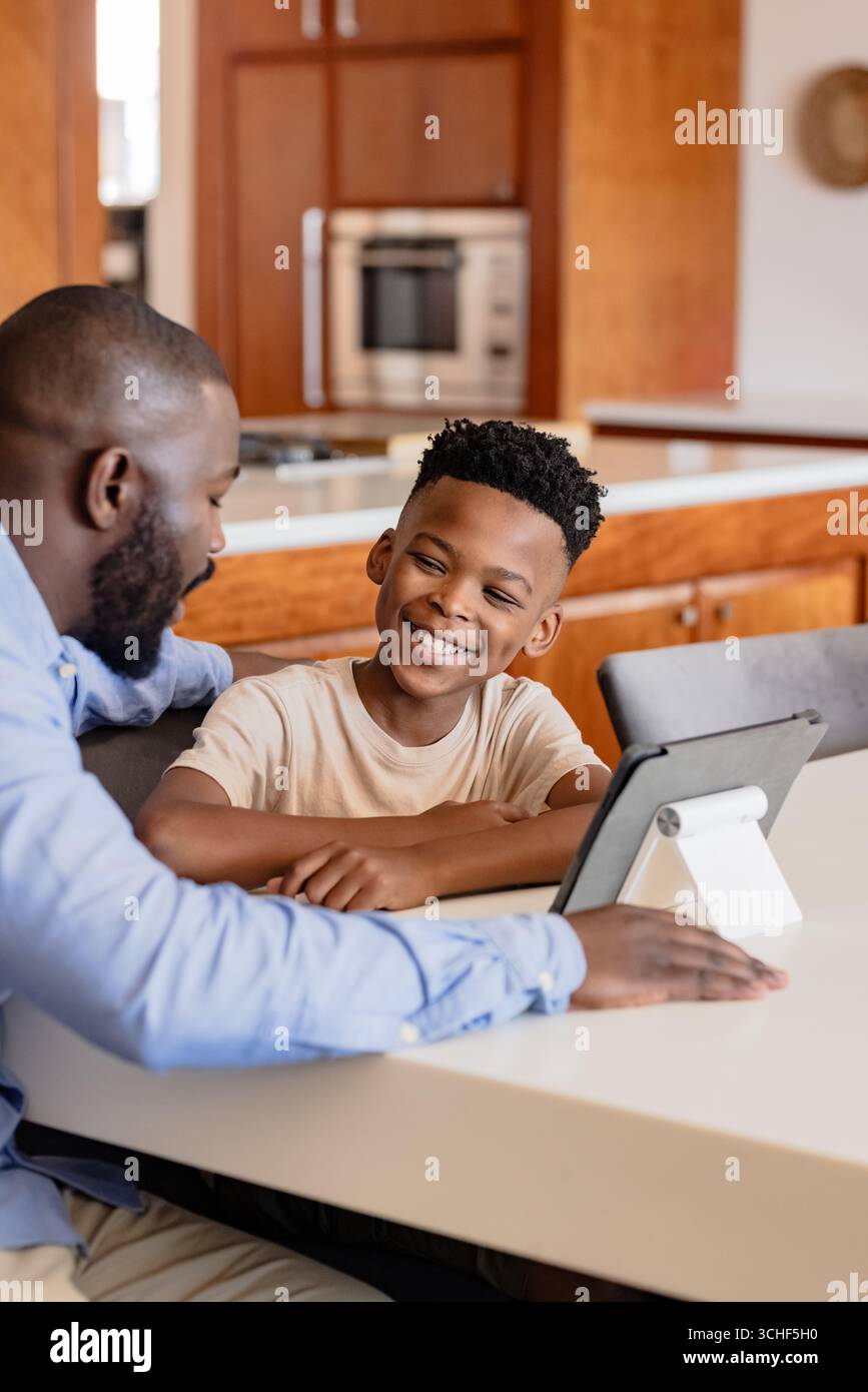Father and son smiling while using tablet together at kitchen table, at home Stock Photo