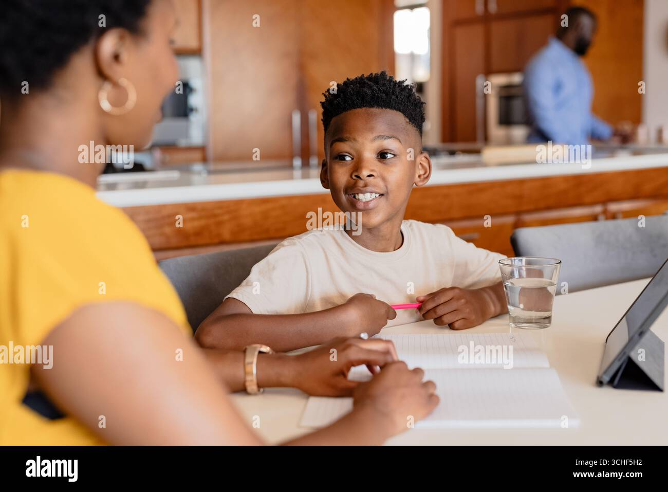 Mother helping son with homework at kitchen table while father prepares ...