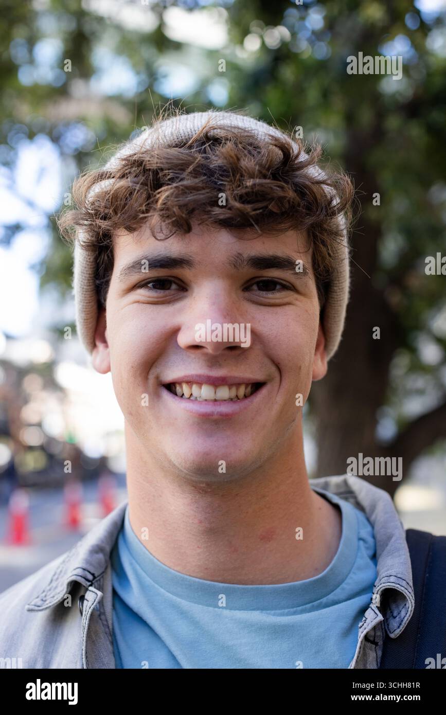 Man wearing brown knit cap, gray overshirt, smiling while standing on sidewalk, backpack strap Stock Photo