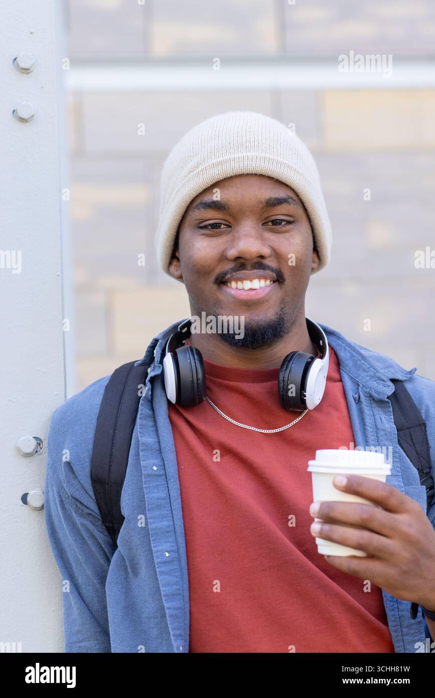 African american man leaning on beam outdoors wearing backpack and headphones with coffee cup Stock Photo