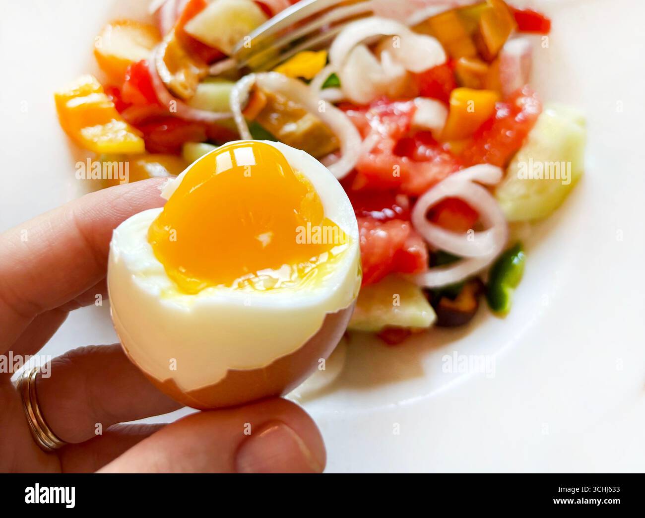 Hand holding a soft-boiled egg, fresh salad in a plate on a white background. Healthy food. Focus on the foreground. Close-up. Stock Photo
