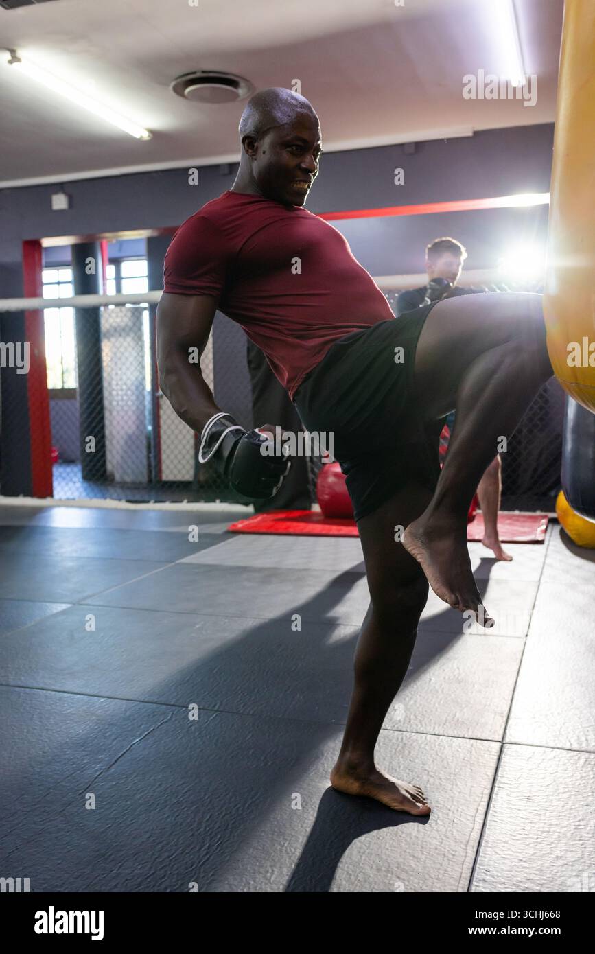 Athlete delivering knee strike to heavy bag in martial arts gym with red, black mats Stock Photo