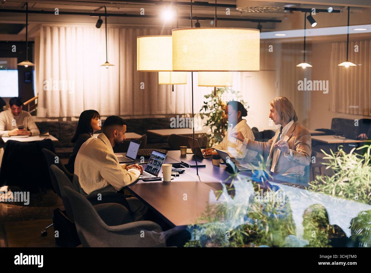 Male and female business colleagues discussing ideas with each other while sitting at desk in illuminated open plan offi Stock Photo