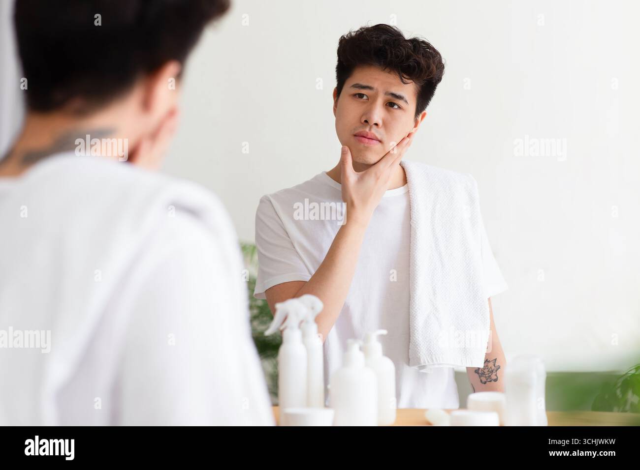Cheerless asian young man with tattoos in white t-shirt, with towel, touches hand to cheek, checks stubble Stock Photo
